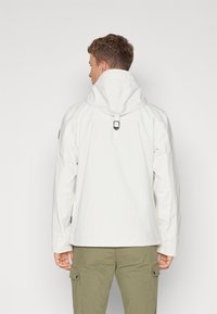 Hvid jakke med en struktureret finish, en hætte og justerbar rem ved halsen. Sammen med grønne cargo bukser med lommer.