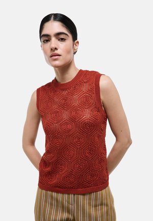 Femme aux cheveux foncés portant un haut sans manches en maille rouge avec un motif hexagonal et un pantalon marron à rayures, debout devant un fond uni.