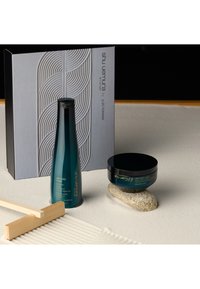 De haarverzorgingsset van Shu Uemura bevat een turquoise glazen shampoo fles en een zwarte pot, beide met een strak ontwerp, geplaatst op een gestructureerd oppervlak.
