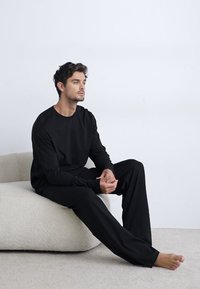 Chemise à manches longues noire et pantalon ample en tissu doux. L'individu est assis sur un canapé texturé de couleur claire.