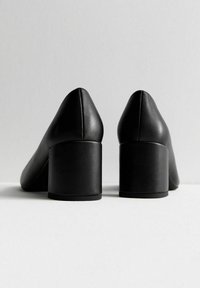 Tacones altos de cuero negro con punta afilada y tacón grueso y cuadrado, con una textura lisa y líneas limpias. Vista trasera mostrada.