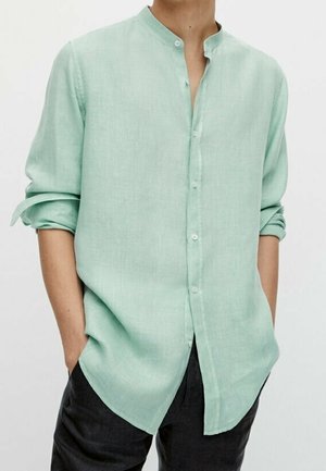 Homme portant une chemise en lin vert clair à col mandarin avec manches retroussées et pantalon noir, mains dans les poches.