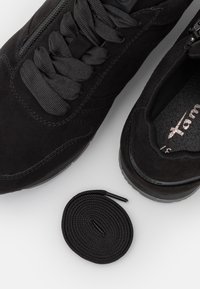 Paire de baskets en daim noir avec de gros lacets noirs, une fermeture éclair latérale et un lacet noir supplémentaire enroulé sur une surface blanche.