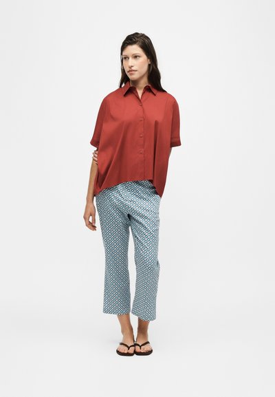 Femme debout, main sur la hanche, portant une chemise rouge ample à manches courtes boutonnée, pantalon court à motifs, et sandales noires sur fond blanc.