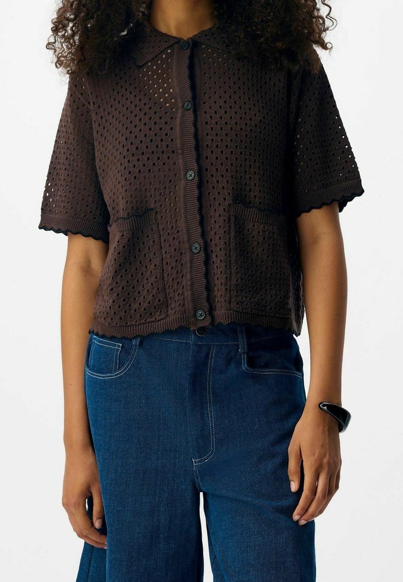 Cardigan marron à manches courtes en maille avec un motif perforé, des bords festonnés et des poches avant. Associé à un pantalon en denim bleu.