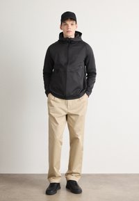 Črna zip-up kapuca s prilegajočim dizajnom, dvema sprednjima žepoma, v kombinaciji s bež chinos hlačami in črnimi supergami, na nevtralnem ozadju.