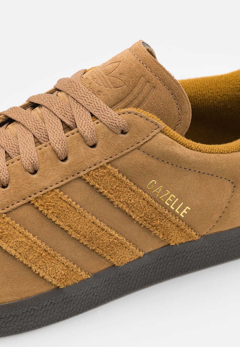 Beżowe zamszowe sneakersy z teksturowanymi paskami, okrągłymi sznurowadłami, złotym logo "Gazelle" oraz czarną gumową podeszwą. Miękka, pluszowa wyściółka wewnętrzna.