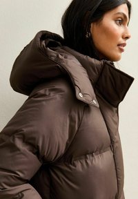 Chaqueta puffer marrón con una gran capucha y cierre de botón a presión, fabricada con un material suave y brillante. Presenta secciones acolchadas para el aislamiento.