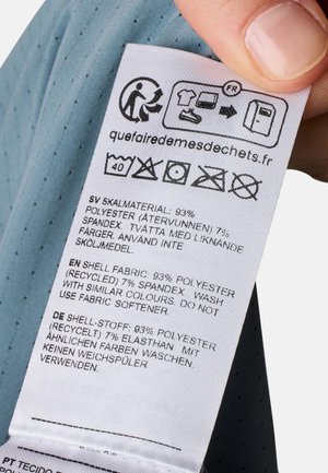 Tøjmærkat holdt i hånden viser vaskebeskrivelser og stofindhold på flere sprog, herunder genanvendt polyester og spandex detaljer.