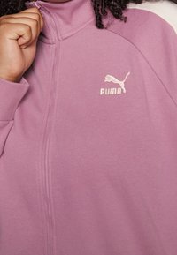 Chaqueta morada con cremallera, confeccionada en tela suave, con cuello alto y un pequeño logo blanco de Puma en el pecho.