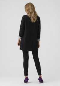 Woman with long blonde hair wears black long blazer over black leggings and purple textured high-heeled shoes, facing away on gray background.