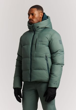Man met groene donsjas met capuchon en zwarte handschoenen, staande tegen een effen lichte achtergrond, kijkt opzij.