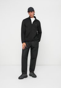 Črna jakna z zadrgo in visokim ovratnikom, v kombinaciji s črnimi trenirkami in črnimi supergami. Gladek material z minimalnimi podrobnostmi blagovne znamke.