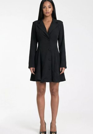 Kobieta ubrana w czarną marynarkową sukienkę zapinaną na guziki z długimi rękawami i czarne wysokie obcasy, stojąca na tle jednolitego białego tła.