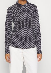 Femme portant un chemisier bleu marine boutonné avec un petit motif géométrique blanc et un pantalon blanc taille haute, se tenant devant un fond uni.