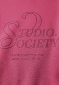 T-shirt in cotone rosa con testo nero: "STUDIO. SOCIETY" e la frase "CREA UNA VITA CHE NON VEDI L'ORA DI INIZIARE."