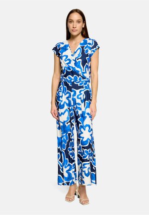 Kobieta ubrana w niebiesko-biały kombinezon z kwiatowym wzorem, z krótkimi rękawami i szerokimi nogawkami, stojąca z rękami swobodnie opuszczonymi wzdłuż ciała.