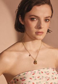 Ung kvinna med kort brunt hår som bär ett axellöst blommigt linne och ett guldsmycke med gröna och rosa ädelstensberlocker.