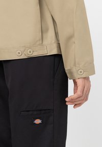 Beige jakke med knapper ved ermene, kombinert med svarte bukser som har lomme og Dickies-logo. Glatt tekstur og skreddersydd design.