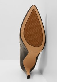 Chaussure à talon haut en cuir noir à bout pointu vue de la semelle, montrant une bande de roulement marron texturée et un talon fin.