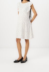 Valkoinen mekko, jossa on mustia pilkkuja, tekstiililtä, jossa on kohokuvioitu pinta, hihaton malli ja levenevä siluetti. Mustat kengät, joissa on koristeellista metalliosaa.