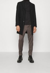 Homme portant un pardessus noir, un pull à col roulé côtelé foncé, un pantalon taupe et des bottines en cuir noir à lacets, debout sur un sol clair.