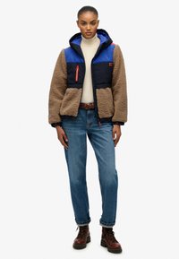 Bruine fleece jack met een blauwe nylon voorkant, voorzien van een zwarte capuchon, een rode ritsaccent en een overall ontspannende pasvorm. Blauwe spijkerbroek en bruine laarzen.
