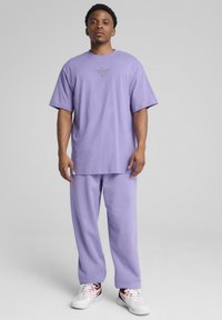 Lilla bomulds-t-shirt med korte ærmer og et logo på brystet, parret med matchende lilla bukser. Hvide sneakers med røde accenter.