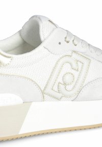 Sneaker bianco con materiali in mesh testurizzati e suede, dettagli dorati e logo in rilievo. Suola spessa e chiara con motivo di trazione.