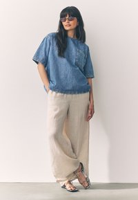 T-shirt oversize in denim con tasca; pantaloni larghi in lino beige chiaro; sandali con motivo argento; occhiali da sole con sottili montature viola.