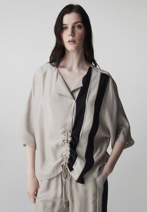 Junge Frau mit dunklen Haaren trägt beiges, locker sitzendes Outfit mit schwarzen und beigen vertikalen Streifen sowie gerafftem Frontdetail, steht vor einfarbigem Hintergrund.