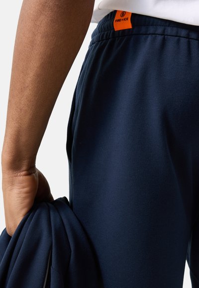 Zbliżenie osoby noszącej granatowe spodnie z pomarańczową metką "Fire+Ice", trzymającej w lewej ręce dopasowany granatowy materiał.