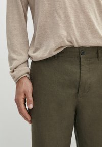 Pantaloni in lino verde oliva con una vestibilità slim, dotati di chiusura a bottone e leggera texture. Una camicia a maniche lunghe di un beige chiaro è parzialmente visibile.
