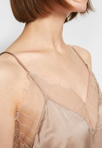 Femme aux cheveux courts et bruns portant un haut en satin beige avec des détails en tissu rayé délicats et transparents près de l'encolure et des fines bretelles.