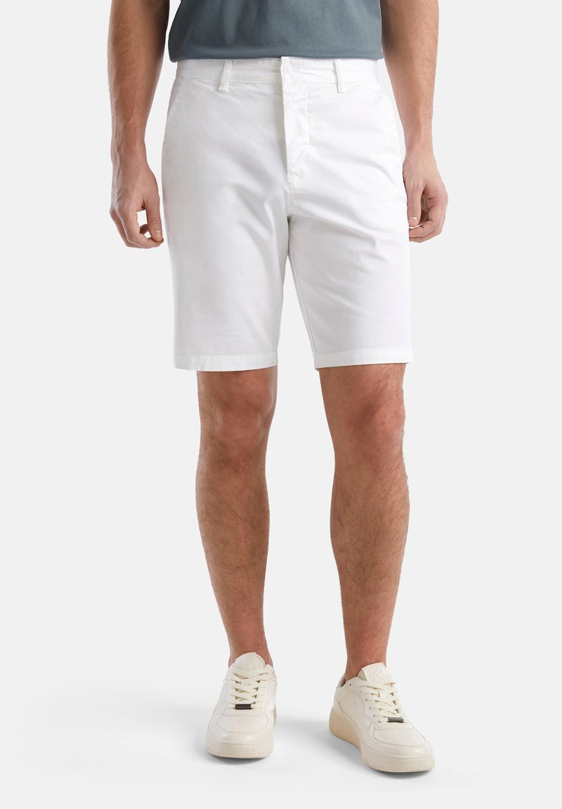 Shorts en coton blanc avec une taille plate, passants de ceinture et poches, associés à des baskets blanches. Le tissu a une texture lisse.
