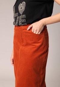 Jupe en velours côtelé orange avec une texture côtelée, dotée de poches latérales et d'une taille cintrée, associée à un t-shirt noir affichant une lettrage orné.