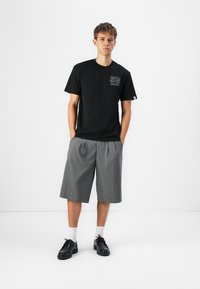 Black cotton t-shirt with logo on chest, paired with knee-length gray pleated shorts and black lace-up shoes. White socks visible.