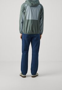 Veste légère à capuche en vert doux et gris, dotée de poignets élastiques et d'une coupe ample. Assortie à un jean denim bleu foncé et des chaussures décontractées.