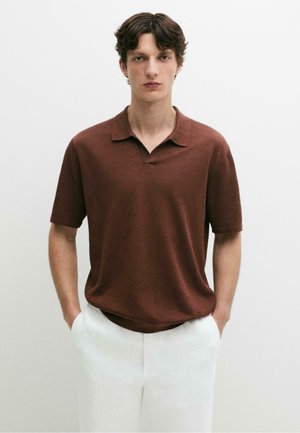 Junger Mann mit kurzen braunen Haaren, der ein braunes kurzärmeliges Poloshirt und weiße Hosen trägt, steht mit den Händen in den Taschen.