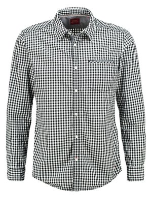 Mężczyzna idzie, ubrany w czarno-białą koszulę w kratę z podwiniętymi rękawami, czarne spodnie, szare tenisówki, trzyma czarne okulary przeciwsłoneczne.