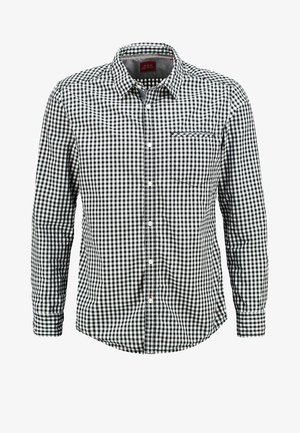 Uomo che cammina indossando una camicia a quadri bianca e nera con le maniche arrotolate, pantaloni neri, scarpe da ginnastica grigie, che tiene degli occhiali da sole neri.