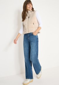 Chaleco acolchado beige con cuello alto, cierre de cremallera y bolsillos laterales, llevado sobre una camisa blanca de manga larga, combinado con vaqueros de pierna ancha azules.