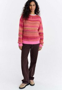 Kobieta o kręconych włosach, ubrana w różowo-pomarańczowy sweter w paski, ciemnobrązowe spodnie, beżowe buty i mały złoty naszyjnik, stojąca na białym tle.