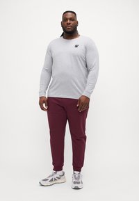 Grå langermet skjorte med en liten svart logo på brystet, paret med maroon joggebukser og grå joggesko med hvite detaljer.