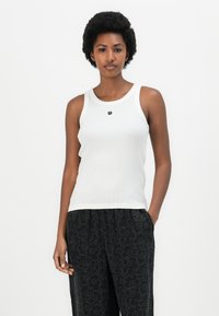 Hvit ermeløs tank topp med rund utrigning, som har et lite svart brodert logo på brystet. Båret med svarte mønstrede joggebukser.