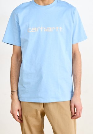 Jasnoniebieska koszulka z krótkim rękawem z beżowym logo "carhartt." noszona przez osobę w beżowych spodniach, z widocznym tatuażem na lewym nadgarstku.