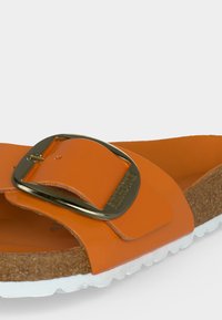 Πορτοκαλί παντόφλα με μεγάλο μεταλλικό αγκράφα, φελλό πάτο και λευκή υφασμένη σόλα.