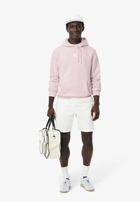 Sudadera rosa claro con bolsillo canguro, pantalones cortos blancos y un bolso blanco con acentos negros. Zapatillas con una mezcla de blanco y gris.