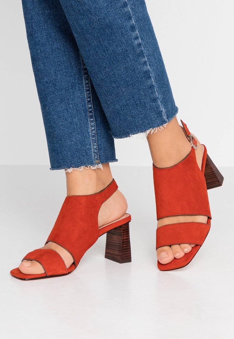 Červené semišové sandály na vysokém podpatku s otevřenou špičkou, nastavovacími řemínky kolem kotníku a texturovaným dřevěným blokovým podpatkem. Nabízí moderní vyřezávaný design.