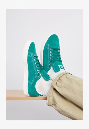 Teal suède sneakers met witte rubberen zolen en veters, voorzien van gestikte accenten en een logo-tag aan de zijkant, gedragen met witte sokken.
